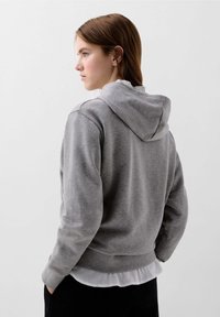 Felpa grigia con cappuccio realizzata in tessuto morbido, caratterizzata da una vestibilità rilassata, polsini a coste e cappuccio con coulisse. Indossata sopra una camicia bianca.