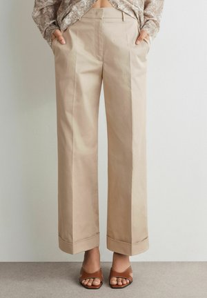 Pantaloni beige a gamba larga con orli risvoltati, indossati con tacchi aperti marroni e una camicia a fantasia infilata. Mani nelle tasche.