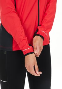 Giacca sportiva rossa con dettagli neri, dotata di polsini elasticizzati e particolari riflettenti. Indossata con leggings neri a trama.