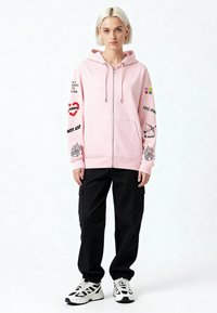 Felpa con cappuccio rosa chiaro con zip e tasca frontale, con stampe grafiche e testo sulle maniche. Abbinata a pantaloni cargo neri e sneakers.