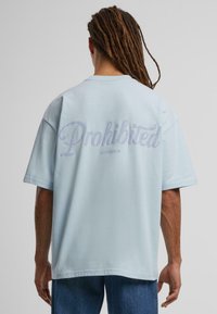 Camiseta de algodón de gran tamaño en azul claro, con la palabra "Prohibido" en cursiva en la parte posterior, y "10119 BERLÍN" debajo.