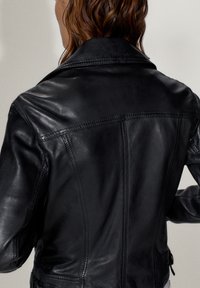 Femme aux cheveux bruns ondulés portant une veste en cuir noire ajustée, vue de dos sur un fond clair uni.