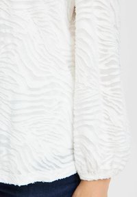 Vit texturerad blus med riplat mönster, genomskinligt material och puffärmar. Sön är ofullbordad med en lätt krullning.