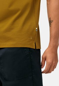Polo giallo senape a maniche corte con spacchi laterali e un piccolo logo. Indossato con pantaloni blu scuro, che mostrano un tessuto testurizzato.