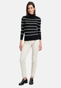 Schwarzer Rollkragenpullover mit weißen horizontalen Streifen, kombiniert mit cremefarbenen schmal geschnittenen Hosen und schwarzen Loafers, stehend vor einem schlichten Hintergrund.