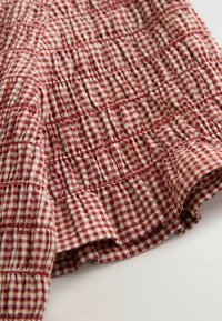 Tessuto a quadretti rossi e crema con trama arricciata, caratterizzato da orlo arruffato e cuciture, che mostra un design leggero e traspirante.