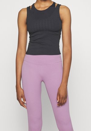 Femme portant un haut noir sans manches ajusté et des leggings taille haute violet clair, debout devant un fond clair uni.