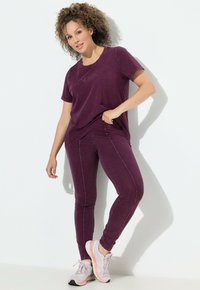 T-shirt bordeaux con testo "sport" e leggings bordeaux abbinati; entrambi offrono una texture morbida. Il modello indossa sneakers grigie chiare con accenti corallo.