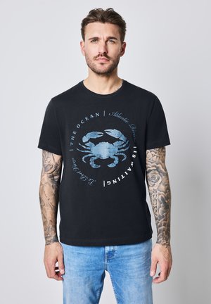 Sort bomulds-t-shirt med et blåt krabbe-grafik og rundt tekst. Kortærmet og rund hals. Modellen har lyseblå jeans på.