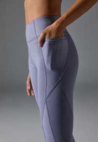 Legginsy sportowe z wysokim stanem w jasnym odcieniu fioletu o gładkiej fakturze, z boczną kieszenią i perforowanymi detalami na dolnej części nogawek.
