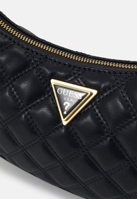 Musta tikattu käsilaukku, valmistettu synteettisestä nahasta, jossa on kullanväriset metalliosat, kolmionmuotoinen logolaatta ja vetoketjusuljenta.