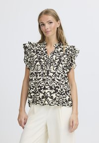 Blusa con motivo floreale nero e crema, con maniche a volant, scollatura a V e vestibilità morbida, abbinata a pantaloni di colore chiaro.