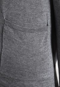 Grått stickat plagg med en framficka med ribbat detalj. Texturen är slät och den övergripande formen är avslappnad och ledig.