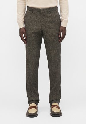 Persona que lleva pantalones texturizados gris oscuro, camiseta crema de manga larga y mocasines marrones y beige, de pie con las manos relajadas a los lados.