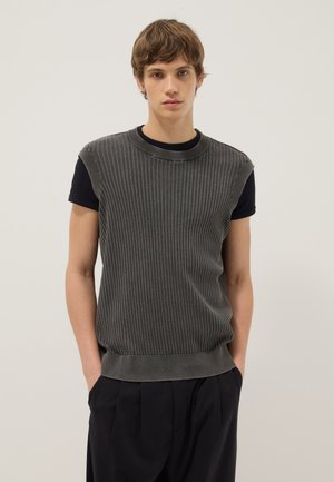 Joven con cabello castaño claro lleva un jersey gris sin mangas de canalé sobre una camiseta negra y pantalones oscuros, de pie frente a un fondo claro y liso.