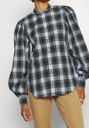 Personne portant une chemise à carreaux noire, blanche et verte à boutons avec des manches longues bouffantes et un pantalon beige.