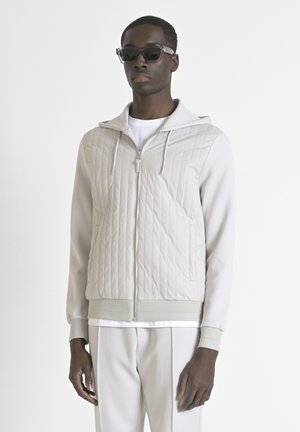 Giacca trapuntata grigia chiaro con cappuccio, cerniera frontale completa e polsini a coste. Abbinata a pantaloni coordinati e una camicia interna bianca.