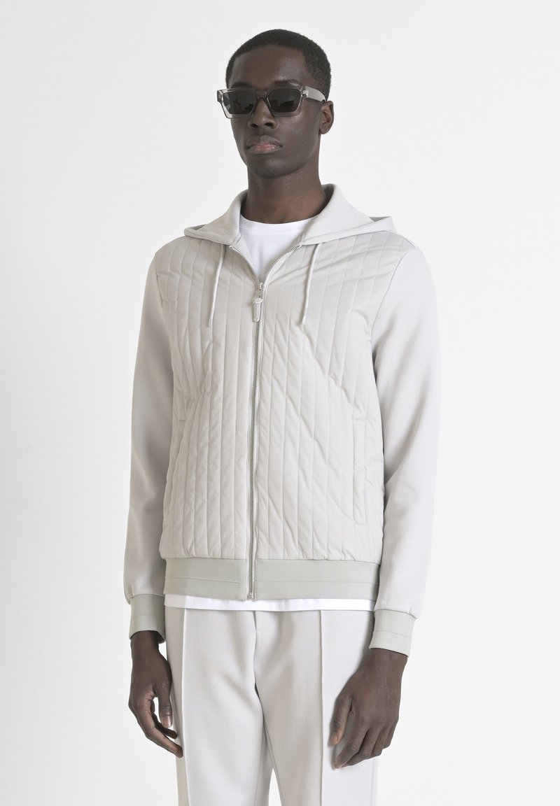 Giacca trapuntata grigia chiaro con cappuccio, cerniera frontale completa e polsini a coste. Abbinata a pantaloni coordinati e una camicia interna bianca.