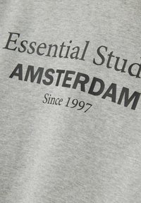 Siva tkanina s crnim tekstom "Essential Stud" djelomično vidljivim, "AMSTERDAM" u podebljanom ikosu, i "Od 1997." ispod.