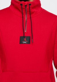 Czerwona polarowa bluza z wysokim kołnierzem, zapinana na zamek z przodu, z czarną naszywką logo Jordana. Posiada sznurki i gładką teksturę.