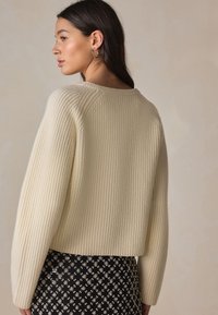 Krémový rebrovaný sveter s okrúhlym výstrihom a spustenými ramenami, v kombinácii s čiernou sukňou so vzorom diamantov a kovovými akcentmi.