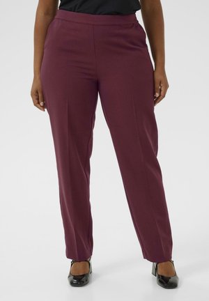 Kvinde iført bordeaux lige ben, høj-taljede bukser med sorte laklæder hæle, stående mod en ensfarvet lys baggrund.