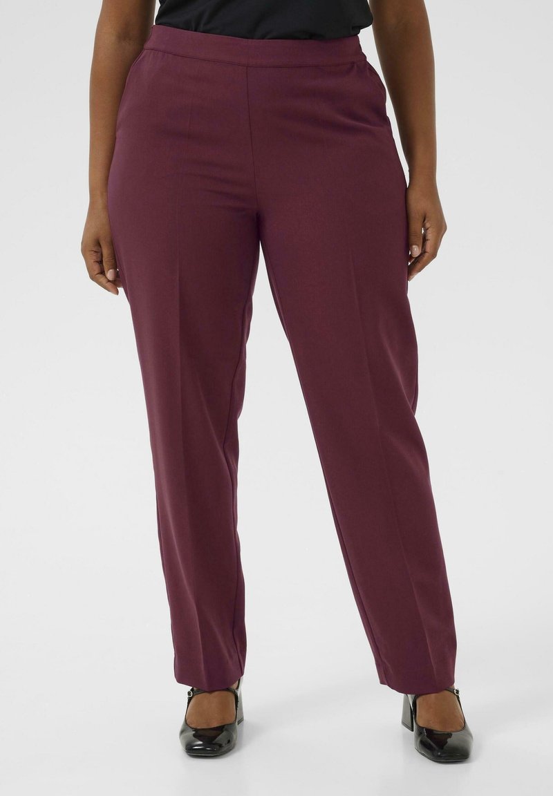 Femme portant un pantalon bordeaux droit à taille haute avec des talons en cuir verni noir, debout devant un fond clair uni.