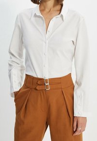 Mujer con una camisa blanca de manga larga abotonada metida dentro de pantalones marrones de cintura alta con detalles de cinturón de doble hebilla.
