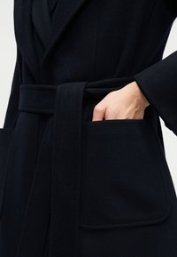 Χέρι που γλιστρά στην μπροστινή τσέπη ενός μαύρου παλτού από μαλλί με ζώνη, φαρδιές πέτλες και μανσέτες στους μανίκες.