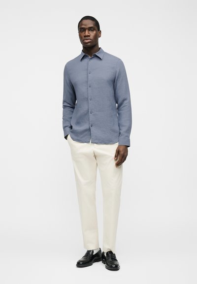 Άνδρας που φοράει μπλε πουκάμισο με μακριά μανίκια και κουμπιά, εκρού παντελόνια και μαύρα μοκασίνια, που στέκεται μπροστά σε απλό λευκό φόντο.