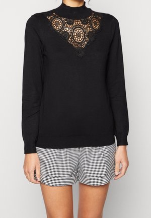 Femme portant un haut noir à manches longues avec un détail en dentelle sur la poitrine et un short pied-de-poule noir et blanc, debout devant un fond uni.