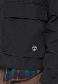 Svart quiltad jacka med en stor framficka och ribbade muddar. Har en liten vit logotyp på fickan. Under jackan finns en rutig skjorta.