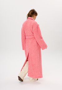 Langer rosa Mantel aus strukturiertem Stoff mit Gürtel in der Taille und seitlichem Schlitz, ausgestattet mit einem Reverskragen und weiten Ärmeln. Trägt man über hellen Hosen.