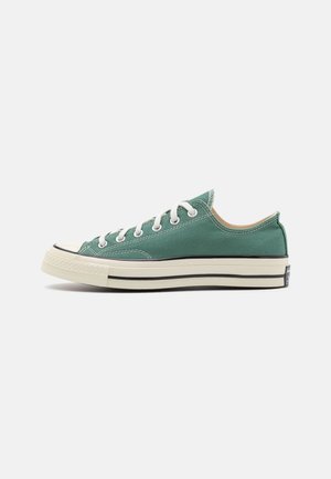 Zapatilla de lona verde baja con cordones blancos y suela de goma con rayas negras, mostrada de lado sobre fondo blanco.
