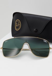 Occhiali da sole Ray-Ban con grandi lenti verdi, montatura in metallo dorato e ponte trasparente. Include una custodia protettiva nera con logo.