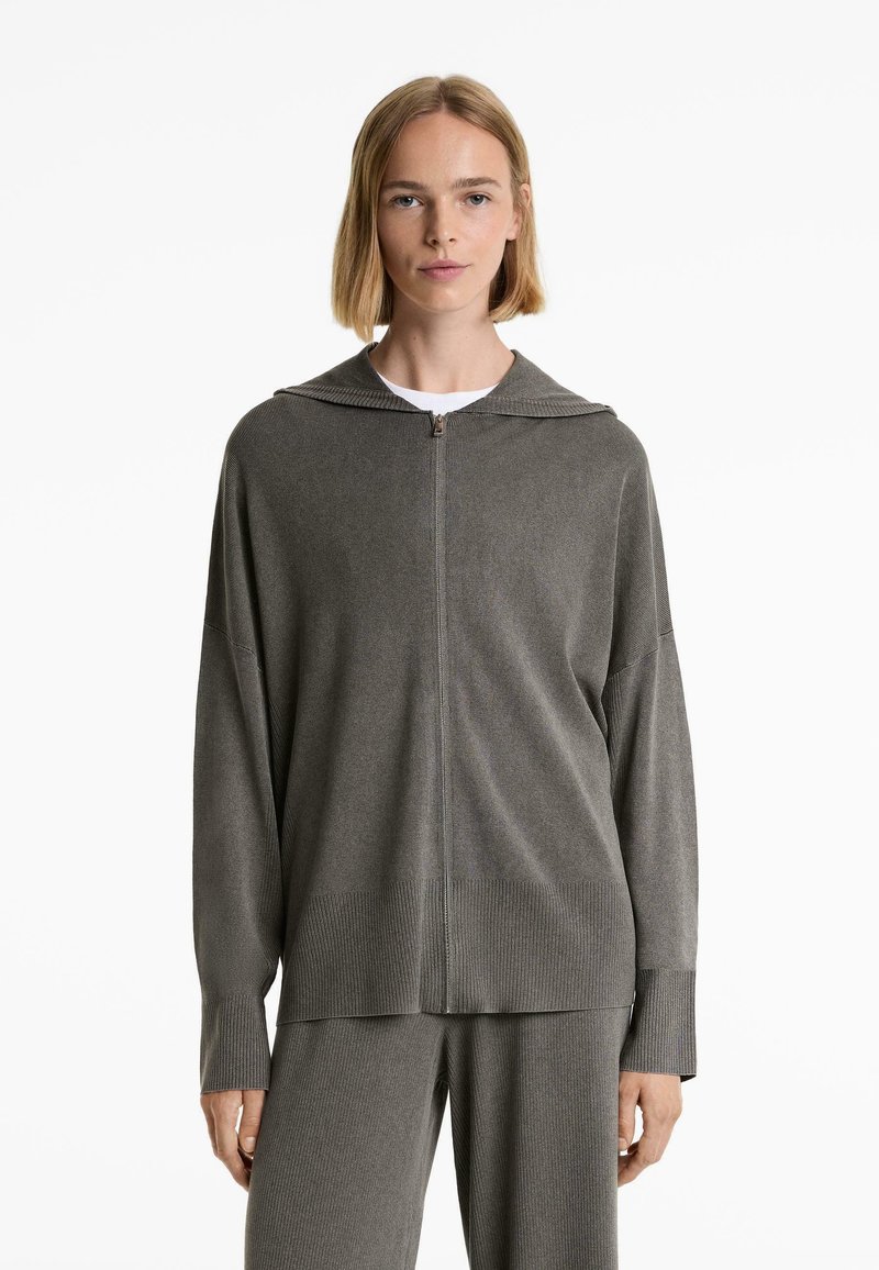 Grå stickad zip-up hoodie med huva, långa ärmar och ribbade mudd; bärs över en vit topp med en avslappnad passform och mjuk textur.