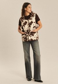 Fleece-Weste mit cremefarbigem Grund und braunem gepunktetem Muster, mit Knopfverschluss und seitlichen Taschen. Getragen über einem schwarzen T-Shirt und grauen Jeans.