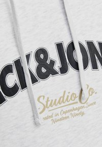 Hellgraue Kapuzenjacke mit dunklem Text in Gold, der "JACK & JONES" und "Studio Co." liest, mit Kordeln und einer weichen Textur.