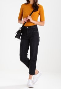 Jeans negros de cintura alta con piernas rectas, combinados con una camiseta de manga corta de canalé mostaza y zapatillas blancas. Bolsa cruzada negra.