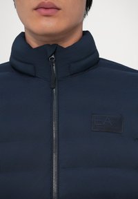 Artimųjų planas, kuriame matyti asmuo, dėvintis tamsiai mėlyną pūkinę striukę su užtrauktuku, pakeltu apykakle ir EA7 Emporio Armani logotipu ant krūtinės.