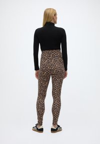 Γυναίκα με την πλάτη, φορώντας μαύρο μακρυμάνικο τοπ, κολάν με λεοπάρ σχέδιο και μαύρα και λευκά αθλητικά παπούτσια σε απλό λευκό φόντο.