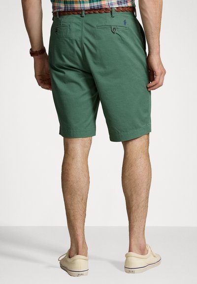 Shorts verzi din bumbac, cu un tiv pliat, având două buzunare la spate și un mic logo. Purtați cu pantofi slip-on din pânză de culoare deschisă.