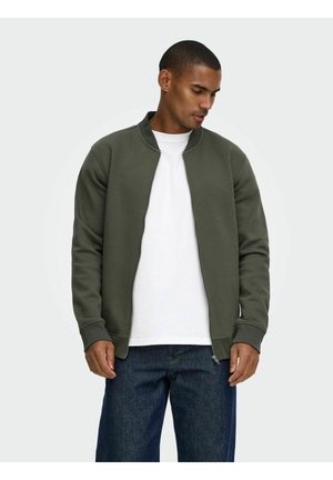 Homme portant une veste zippée vert foncé par-dessus une chemise blanche et un jean bleu foncé, regardant vers le bas et sur le côté sur un fond uni.