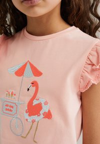 Camiseta de algodón rosa con mangas con volantes. Presenta un bordado colorido de un flamenco, una flotador y un carrito de helados con una sombrilla a rayas.