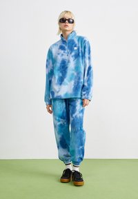 Fleece genser og bukser i blå tie-dye mønster. Har en glidelåskrage, elastiske mansjetter og ribbede detaljer. Kombinert med svarte joggesko.