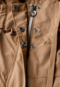 Jasnobrązowa kurtka z tkaniny z zamkiem z przodu, srebrnymi elementami, zatrzaskami i teksturowanym wykończeniem. Zawiera regulowane paski.