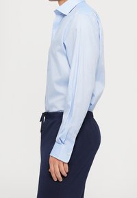 Man med ljusblå långärmad skjorta med knappar, instoppad i mörkblå byxor med elastisk midja, sedd från sidan mot en enkel bakgrund.