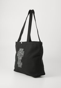 Black canvas tote bag with two shoulder straps, featuring white embroidered city names arranged vertically on one side.