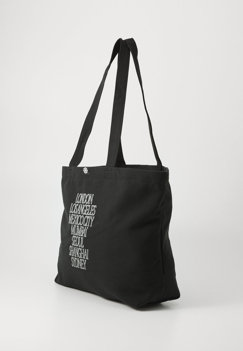 Sac fourre-tout noir en toile avec deux poignées, présentant des noms de villes brodés en blanc disposés verticalement sur un côté.