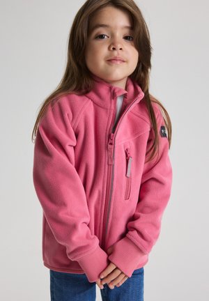 Junges Mädchen mit langen braunen Haaren, trägt eine rosa Fleecejacke mit Reißverschluss und blaue Jeans, steht mit verschränkten Händen vor sich.